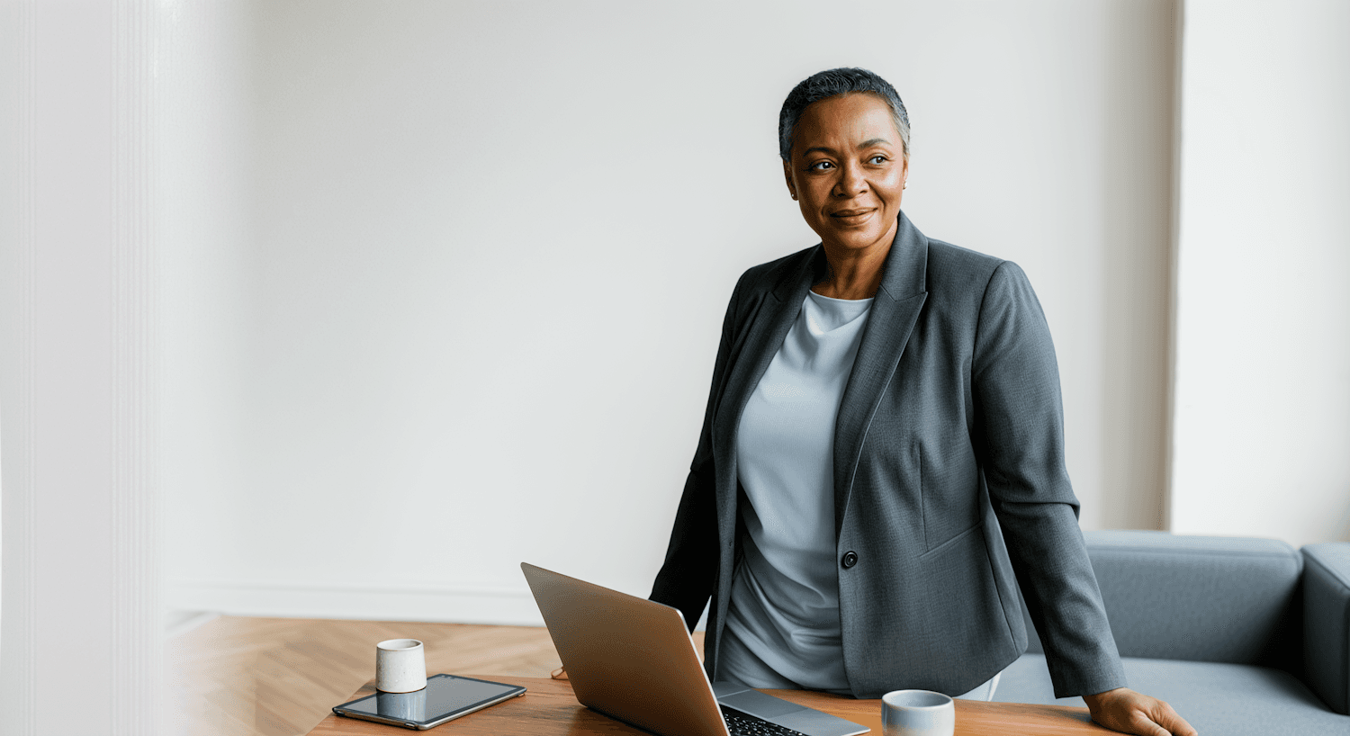 Woman in a tailored grey blazer and pale blue top standing at a desk with a laptop